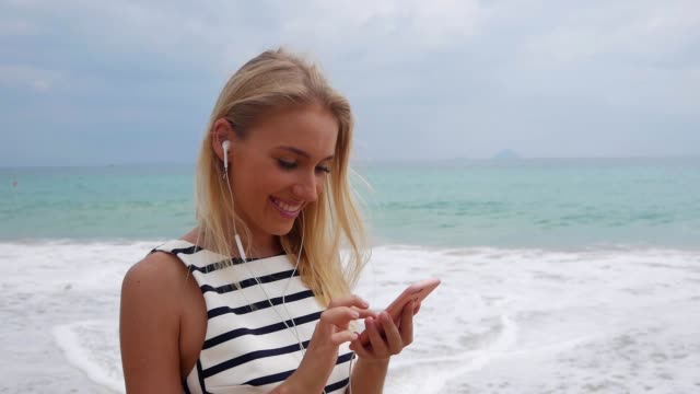 Young-beautiful-slim-woman-with-long-blonde-hair-in-black-and-white-dress-standing-on-the-coast-and-using-smartphone-over-background-at-storm-on-the-sea..-Girl-on-the-beach-touching-screen-and-smile.