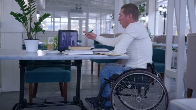 Online-Training,-Studenten,-Behinderte-im-Rollstuhl-Notizen-im-Notizbuch-während-Fernausbildung-mit-Laptop-Computer-im-Café-am-Tisch-sitzen