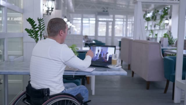 Home-education,-disabled-man-in-wheelchair-writes-notes-in-notebook-while-watching-online-training-on-laptop-computer-sitting-at-a-table-in-restaurant