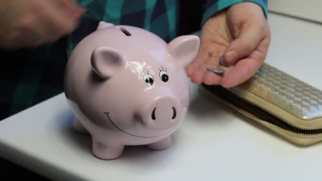 On-the-table-there-is-a-piggy-bank-in-the-form-of-a-pink-pig.-Lying-wallet-and-scattered-coins.-A-man-collects-coins-from-the-table,-puts-them-in-a-piggy-bank-and-closes-the-wallet.