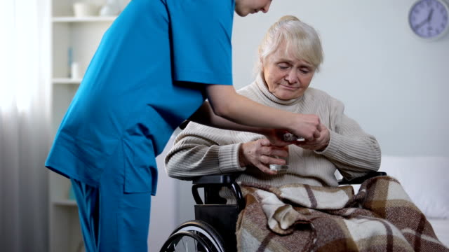 Junge-Krankenschwester-geben-Medikamente,-alte-Dame-im-Rollstuhl,-Reha-Zentrum