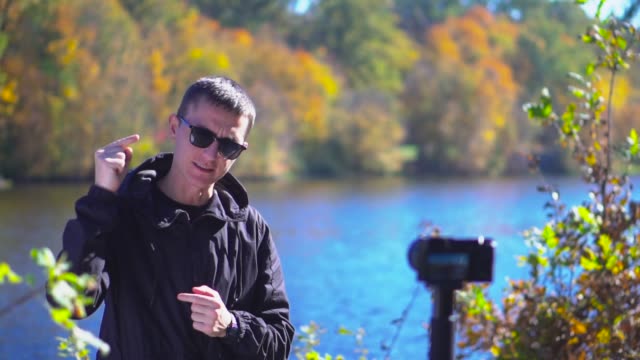 Blogging-young-man-in-sunglasses-stand-next-to-camera-at-nature-showing-thumbs-up-and-saying-goodbye-for-subscribers-slow-motion