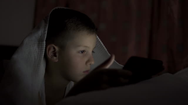 Ein-fröhlicher-Junge-liegt-im-Bett-unter-einer-Decke-und-spielt-auf-einem-Tablet-in-einem-Spiel-im-Dunkeln.-Das-Gesicht-des-Kindes-wird-von-einem-hellen-Monitor-beleuchtet