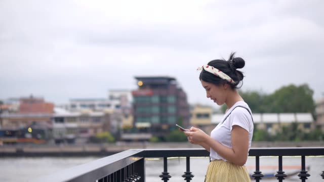 Hermosa-joven-asiática-usando-el-teléfono-inteligente-disfrutando-de-leer-las-redes-sociales-al-lado-de-la-valla-en-el-río.