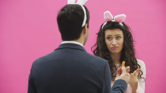 Beautiful-young-girl-standing-on-a-pink-background.-During-this,-there-are-ears-of-rabbits-on-the-head.-A-man-stands-back-to-the-camera,-holding-a-carrot-showing-the-girl.-The-girl-does-not-like-carrots,-leaving-the-frame.-The-man-looks-in-the-frame.