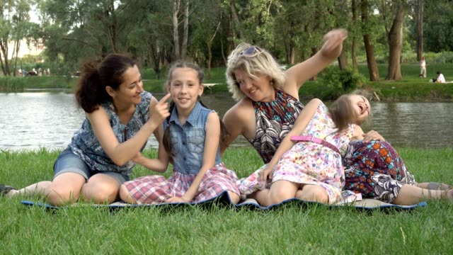 Glückliche-gleichgeschlechtliche-Familie:-Zwei-Mütter-und-zwei-kleine-Töchter-spielen-und-haben-Spaß-am-Sitzen-auf-dem-Rasen-im-Park-bei-Sonnenuntergang-an-einem-Sommertag-gegen-den-Fluss.-Familienurlaub-in-der-Natur.-4K.
