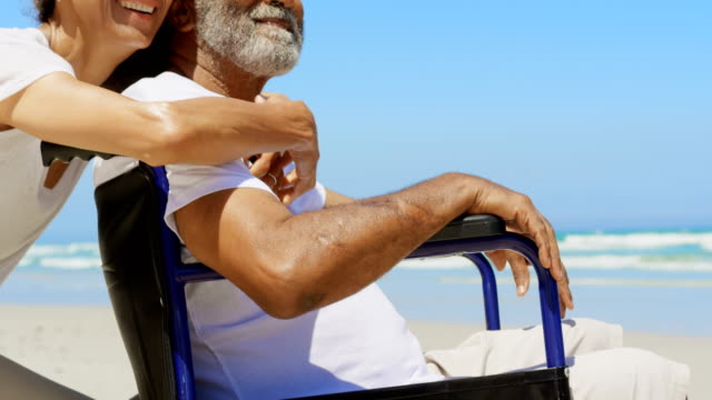 Side-view-of-active-senior-African-American-woman-embracing-disabled-senior-man-on-the-beach-4k