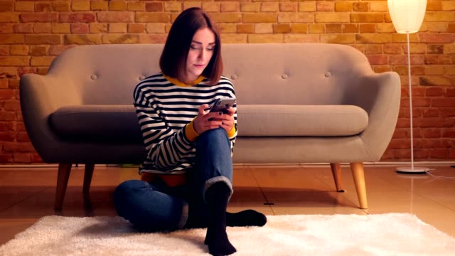 Closeup-portrait-of-young-pretty-girl-using-the-phone-sitting-on-the-floor-in-a-cozy-apartment-indoors