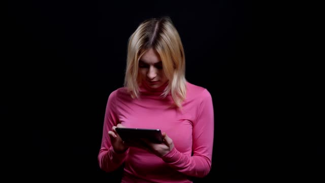 Closeup-shoot-of-young-pretty-blonde-female-using-social-media-on-the-tablet-in-front-of-the-camera-with-background-isolated-on-black