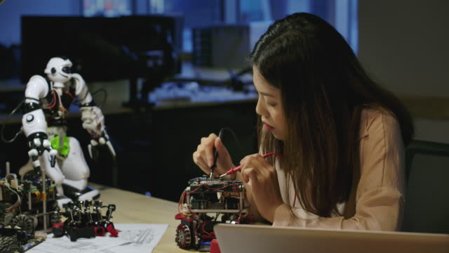 Las-jóvenes-ingenieras-de-desarrollo-electrónico-asiático-trabajan-con-el-robot,-midiendo-la-señal-en-los-circuitos-eléctricos-del-prototipo-de-robótica-en-el-taller.-Personas-con-concepto-de-tecnología-o-innovación.
