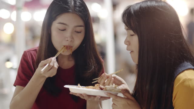 Asian-young-women-friends-travel-in-Bangkok,-Thailand,-beautiful-female-feeling-happy-walking-and-eating-Pad-Thai-at-The-Khao-San-Road.-Women-travel-eat-street-food-concept.-Slow-motion-shot.