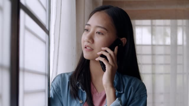 Porträt-lächelnde-junge-asiatische-Frau-am-Telefon-mit-Freunden,-die-neben-dem-Fenster-im-Home-Office-stehen.