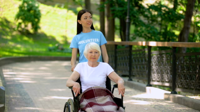 Happy-social-volunteer-walking-hospital-garden-with-disabled-elderly-pensioner