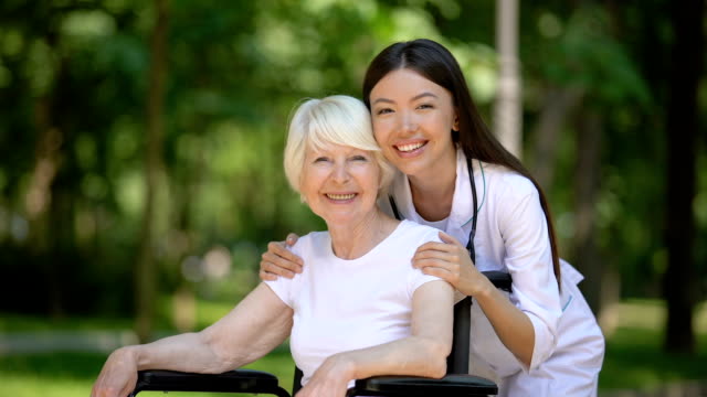 Lächelnde-Weibliche-Krankenschwester-umarmt-behinderte-ältere-Frau-und-Blick-in-die-Kamera