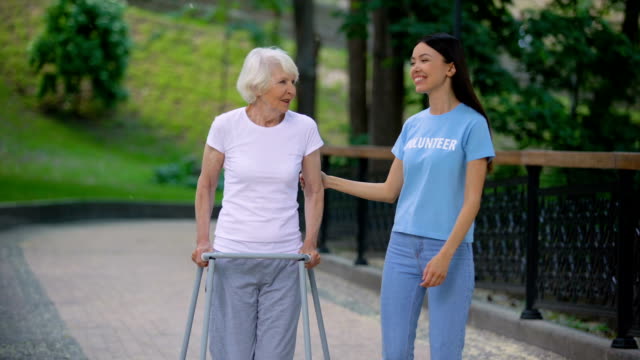 Caring-young-woman-supporting-old-lady-with-walking-frame,-social-volunteering