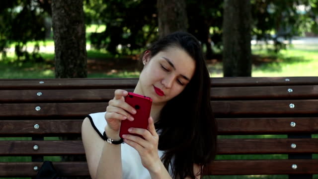Nahaufnahme-Porträt-Ein-schönes-junges-Mädchen-in-einem-weißen-T-Shirt-plaudert-in-sozialen-Netzwerken-auf-ihrem-Smartphone,-während-sie-auf-einer-Bank-in-einem-Park-sitzt.