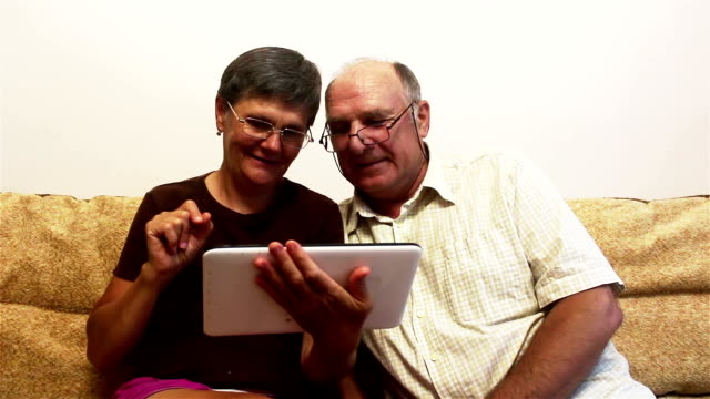 Ein-älteres-Ehepaar-sitzt-zu-Hause-auf-einem-Sofa-und-schaut-sich-Fotos-auf-einem-Tablet-PC-an,-lächelt-und-redet-aktiv.