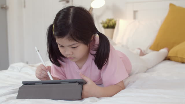 Young-Asian-girl-drawing-at-home.-Asia-japanese-woman-child-kid-relax-rest-fun-happy-using-tablet-draw-cartoon-before-sleep-lying-on-bed,-feel-comfort-and-calm-in-bedroom-at-night-concept.-Slow-motion