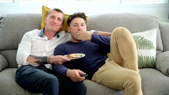 Schwules-Paar-entspannen-doneriert-auf-der-Couch-und-schaut-fern.-Kekse-essen-und-lachen.