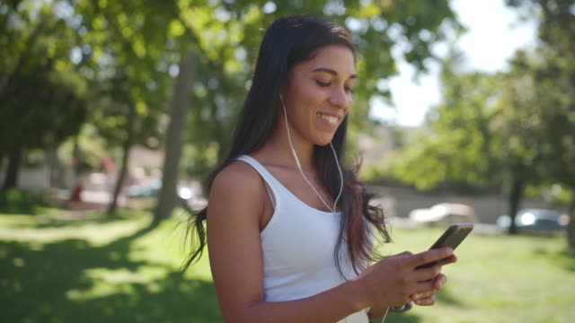Fröhliche-junge-Brünette-gemischte-Rasse-Frau-mit-Kopfhörern-in-den-Ohren-schauen-in-die-Kamera-und-lächelt-weiße-SMS-Nachrichten-auf-Handy-im-Park