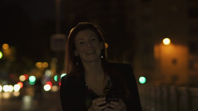 Caucasian-modern-woman-wearing-flower-dress,-black-jacket-and-red-hair-walking-through-the-street-and-writing-a-text-message-on-smartphone-and-laughing-at-camera-by-night.-Paris-4K-UHD.-Slow-Motion.