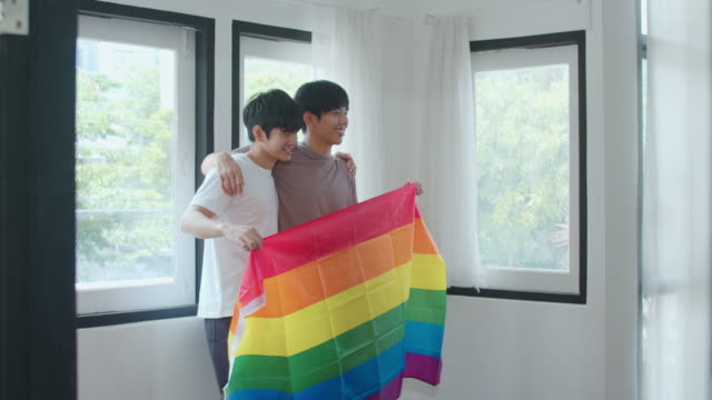 Porträt-Junge-asiatische-Homosexuell-Paar-Gefühl-glücklich-zeigt-Regenbogen-Flagge-zu-Hause.-Asia-LGBTQ+-Männer-entspannen-zu-schüchtern-Lächeln-blickend-in-die-Kamera,-während-Umarmung-im-modernen-Wohnzimmer-im-Haus-im-MorgenKonzept.