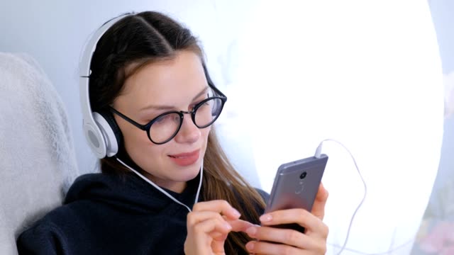 Frau-hört-Musik-in-Kopfhörern-auf-dem-Smartphone-sitzen-im-Sessel-zu-Hause.