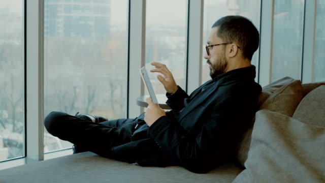Junger-Geschäftsmann-sitzt-auf-der-Couch-und-benutzt-ein-digitales-Tablet.-4K