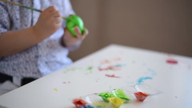 The-hands-of-a-litlle-child-with-a-brush-paint-an-Easter-egg-in-green-color-on-the-table.