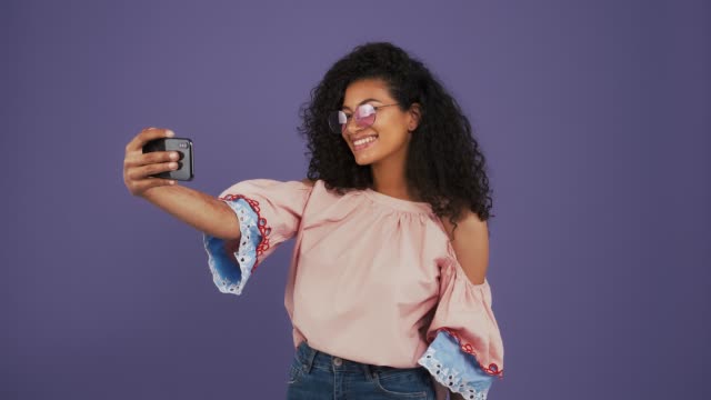 Afro-american-lady-in-casual-clothes.-She-enjoying-video-call-on-her-smartphone,-smiling,-sending-air-kiss.-Posing-on-purple-background.-Close-up