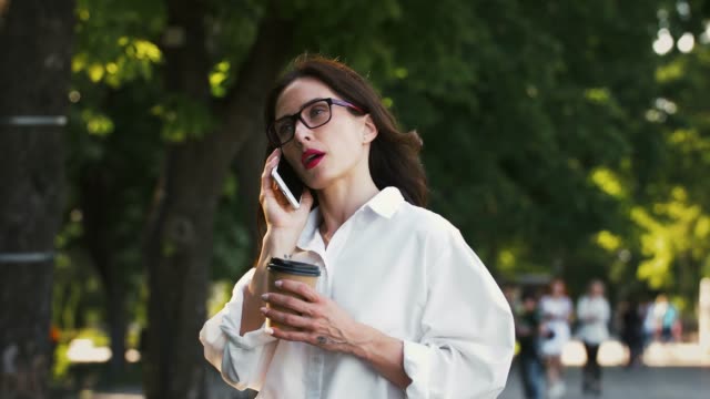 Weiblich-in-Brille,-weißes-Hemd.-Sprechen-auf-dem-Smartphone-und-halten-Tasse-Kaffee,-stehen-deiner-Park-mit-grünen-Bäumen.-Geschäftskonzept.-Zeitlupe