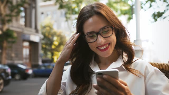 Business-girl-in-glasses,-white-shirt.-Looking-surprised-and-smiling,-reading-news-using-smartphone.-Sitting-in-outdoor-cafe.-Close-up,-slow-motion