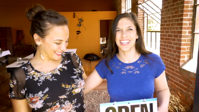 Portrait-of-two-beautiful-female-business-partners-hold-an-open-sign-and-smile