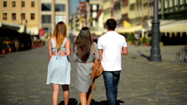 Rückansicht-von-drei-Freunden-Kaffee-zu-trinken-und-zu-Fuß-durch-die-Stadt.-Zwei-Mädchen,-die-tragen-Sonnenbrillen-und-kurze-Kleider-und-hübscher-Junge-im-weißen-Hemd-und-Jeans,-Zeit-miteinander-zu-verbringen