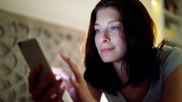 Close-up-of-young-smiling-woman-using-smartphone-lying-in-bed-at-home-at-night
