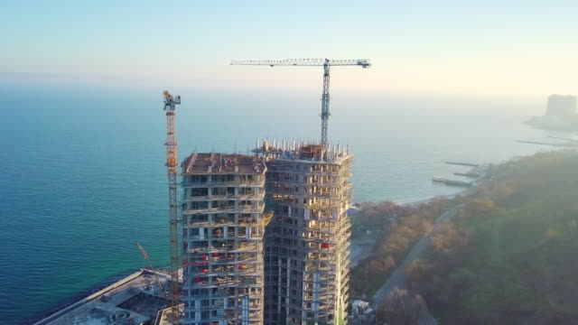 Aerial-city-view.-Construction-of-a-high-rise-skyscraper-on-the-ocean-by-two-cranes.-The-camera-moves-horizontally.-Lower-shooting-height