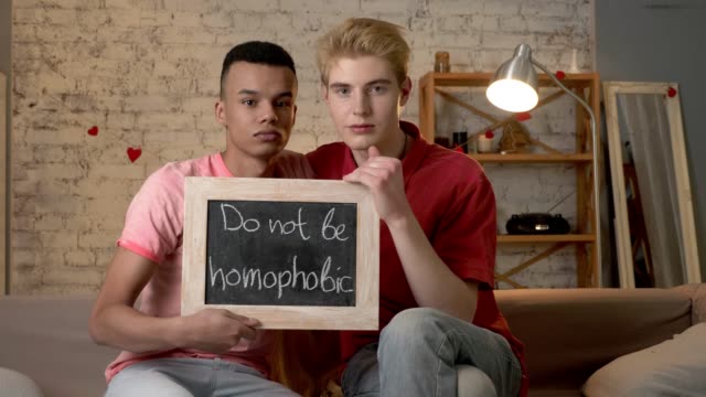 A-sad-international-gay-couple-is-sitting-on-the-couch-and-holding-a-sign.-Do-not-be-homophobic.-Look-at-the-camera.-Home-comfort-on-the-background.-60-fps