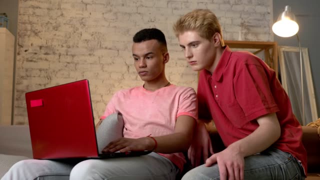 Two-multinational-homosexual-friends-sit-on-the-couch,-use-a-laptop,-watch-an-interesting-video-on-the-Internet,-surprised.-Home-cosiness,-family,-internet-concept.-60-fps