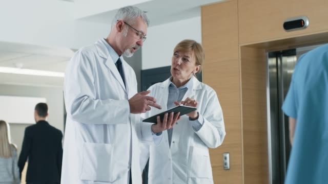 In-the-Hospital-Doctors-have-Discussion-while-Using-Tablet-Computer.-In-the-Background-Patients-and-Medical-Personnel.-New-Modern-Fully-Functional-Medical-Facility.