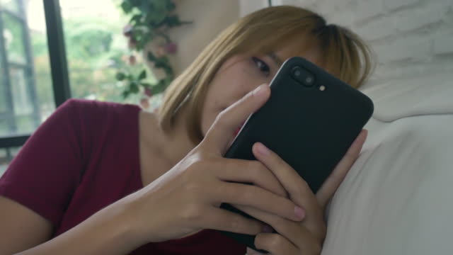 Mujer-asiática-jugando-smartphone-mientras-está-acostado-en-el-sofá-de-casa-en-su-sala-de-estar.-Feliz-mujer-utilice-el-teléfono-para-mensajes-de-texto,-lectura,-mensajería-y-compra-en-línea-en-casa.-Mujer-de-estilo-de-vida-al-concepto-de-hogar.