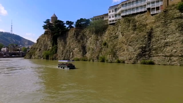 Touristen-auf-Boot-schwimmt-auf-Fluss-Kura,-Tiflis,-Metekhi-Kirche-im-Hintergrund
