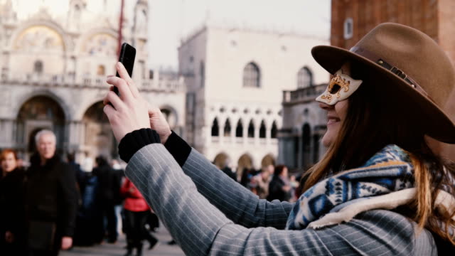 Glückliche-Frau-mit-einem-stylischen-Hut-und-weißen-Karnevalsmaske-nimmt-Selfie-Foto-lächelnd-am-Stadtplatz-von-Venedig,-Italien.