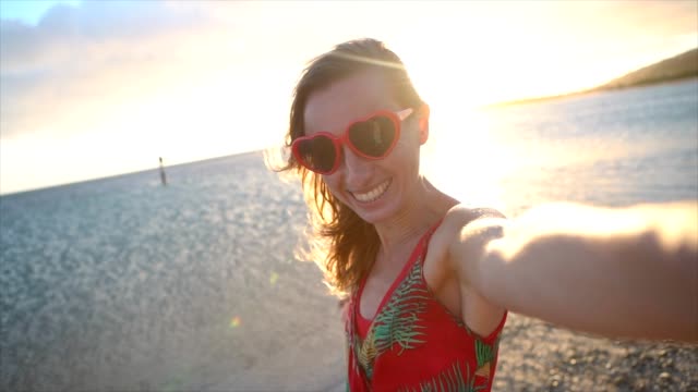 Mujer-joven-con-el-corazón-en-forma-de-gafas-de-sol-rojo-tomar-selfie-fotografía-al-atardecer-en-la-hermosa-playa-en-Hawai.-Cámara-lenta