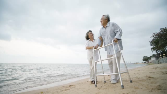 Senior-man-exercise-with-walker-and-his-wife-assist-him-walking-along-beach-in-slow-motion.-People-with-healthcare,-medical-and-retirement-concept.