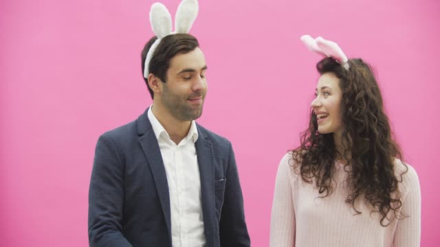 Young-beautiful-couple-standing-on-a-pink-background.-With-hackneyed-ears-on-the-head.-During-this-man-gives-his-wife-a-basket-of-colored-eggs.