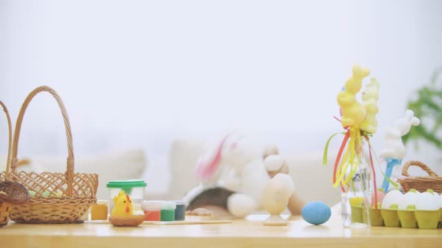 Little-boy-is-hiding-under-the-wooden-table,-full-of-Easter-decorations:-basket,-yellow-chicken,-colorful-eggs-pains-and-paint-brush.-Boy-is-playing-with-a-cute,-soft-white-bunny-with-pink-ears,-on-the-table.