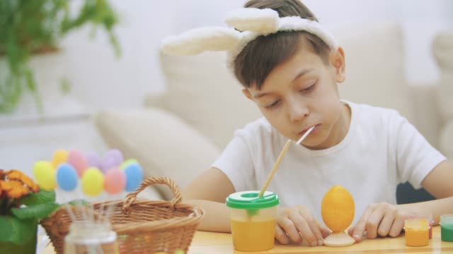 Intersted-and-concentrated-boy-is-finishing-to-colourize-an-Easter-egg-in-an-yellow-colour,-sitting-at-the-desk-and-tasting-sweet-lollipop-and-showing-his-right-thumb-up.