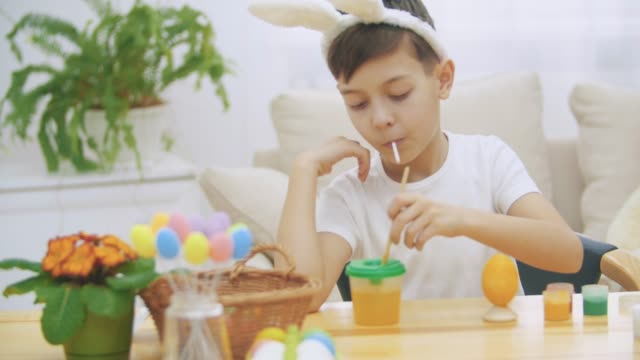Intersted-and-concentrated-boy-is-finishing-to-colourize-an-Easter-egg-in-an-yellow-colour,-sitting-at-the-desk-and-tasting-sweet-lollipop-and-showing-his-thumbs-up.
