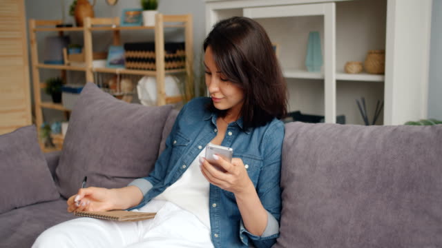 Smart-woman-writing-in-notebook-using-smartphone-smiling-sitting-on-sofa-at-home