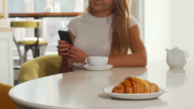 Frau-mit-Smartphone-im-Bäckerei-Café,-leckeres-Croissant-im-Vordergrund